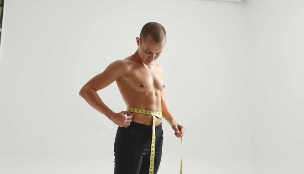 A well-lit, high-resolution image of a person standing in a clean, minimalist studio space, carefully measuring their body with a soft tape measure. The subject's face is not visible, maintaining a focus on the measurement process. The lighting is bright, natural, and evenly distributed, creating a sense of clarity and precision. The camera angle is positioned at waist level, capturing the measurement process from an informative, instructional perspective. The background is a simple, uncluttered white or light gray, allowing the subject and the measurement activity to be the central focus of the image.